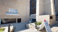 This single-floor commercial space is currently available for lease and is housed within a substantial concrete building with a distinctive brutalist architectural aesthetic. The exterior features textured concrete panels, large dark-framed windows with blue accents, and a paved entry area with accessible ramp access and landscaped planters flanking the entrance.

Inside, the space transitions into a well-appointed fitness facility. The main workout area is generously sized, featuring durable rubber-speckled flooring designed to withstand heavy use. Exposed structural ceiling elements are painted white and fitted with rows of bright fluorescent lighting, contributing to an open and energetic atmosphere. Large windows along one wall flood the space with natural light while offering views to the exterior. Full-length mirrors line one wall, enhancing the sense of space and supporting functional training needs.

The gym is equipped with cable machines, rowing equipment, free weights, benches, and a dedicated cardio section featuring treadmills and upright bikes, each positioned beneath wall-mounted flat-screen televisions. A corridor with tile flooring and drop-panel ceilings connects to additional rooms, including a separate entry door, offering flexibility for varied commercial or wellness-oriented uses.