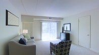 This single-floor residential space offers a comfortable and well-appointed living environment designed with functionality and natural light in mind. The living area serves as the central gathering space, featuring soft wall-to-wall carpeting that adds warmth underfoot. Large vertical blinds frame a full-width sliding glass door, flooding the room with abundant natural light and providing a seamless connection to the outdoors, where lush greenery is visible beyond. The neutral color palette of soft whites and warm grays creates a calm, versatile backdrop suited to a variety of furnishing styles. A louvered interior door adds a classic architectural detail, while ceiling vents indicate a centralized HVAC system for year-round climate comfort. The space accommodates a thoughtful furniture arrangement, with room for multiple seating options that invite relaxed conversation or quiet reading. A dark wood dresser with a framed mirror anchors one wall, providing both storage and visual interest. The overall ambiance is airy and understated, with clean lines and an open feel that makes the space feel welcoming and move-in ready. The layout flows naturally, offering a practical and pleasant living environment.