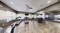 The fitness center at 2002 Battlecreek Dr is a thoughtfully designed exercise facility offering a comprehensive workout experience across a single floor. The space is divided into two functional zones, providing dedicated areas for both cardio training and strength or flexibility work.

The main exercise area features rich hardwood-style flooring, two-tone dark and light gray walls with crown molding, and recessed lighting complemented by ceiling fans for optimal air circulation. Multiple windows fitted with blinds allow generous natural light to fill the space while maintaining privacy. Wall-mounted flat-screen televisions are positioned for convenient viewing during workouts.

The cardio zone is well-appointed with multiple treadmills, an elliptical cross-trainer, and a stationary cycle, offering a variety of aerobic options. A cable crossover machine and a tiered dumbbell rack support strength training needs. The secondary room features rowing machines, stability balls on a dedicated rack, an adjustable workout bench, and floor mats, making it ideal for functional training and flexibility exercises. Full-length mirrors line the walls of both areas, enhancing the sense of space and allowing users to monitor their form. French doors provide a welcoming entry point, rounding out this well-equipped community fitness facility.