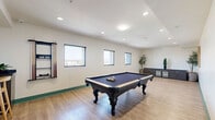 Located in Escondido, CA, this well-appointed game room offers a thoughtfully designed space for recreation and socializing. The room is anchored by a classic full-size pool table with an elegant dark felt surface and ornate carved legs, accompanied by a wall-mounted cue rack that keeps equipment neatly organized and accessible.

Light wood-look flooring runs throughout, lending the space a clean, modern feel that is both inviting and easy to maintain. Teal baseboards provide a vibrant accent that adds a touch of personality to the otherwise neutral white walls. Recessed lighting and flush-mount ceiling fixtures ensure the space is well-lit throughout the day and evening.

A built-in bar-height counter along one wall is paired with wooden bar stools, creating a comfortable area for spectators and casual gatherings. Nearby, a dark low-profile storage credenza with glass-front cabinet doors provides additional functionality, while potted greenery adds a refreshing natural element. A framed decorative artwork piece on the wall ties the room's aesthetic together with a nod to local Southern California culture.

Multiple windows along the exterior wall allow natural light to fill the room and offer views of the surrounding area. A connecting hallway with teal-trimmed doorways provides convenient access to the broader facility. This game room is a versatile and engaging amenity space.