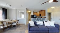 This 2-bedroom, 2-bathroom single-floor residence offers a well-designed open-concept layout that seamlessly connects the living, dining, and kitchen areas. The living room serves as the centerpiece of the home, anchored by wood-look flooring that extends throughout the main living areas, lending warmth and continuity to the space. Light gray walls provide a neutral backdrop that enhances the sense of openness and brightness.

The kitchen is outfitted with warm-toned wood cabinetry, black appliances, and a breakfast bar with seating, making it both functional and inviting. Track lighting above the kitchen adds focused illumination, while the open pass-through design keeps the space feeling connected to the rest of the home.

Adjacent to the living area, the dining space accommodates a table and chairs, complemented by a wall-mounted mirror that reflects natural light and adds depth to the room. A ceiling fan is visible overhead, contributing to the comfortable climate of the space.

Hallway access leads to the two bedrooms, each offering privacy from the main living areas. Bi-fold closet doors are noted, maximizing storage efficiency. Overall, the layout is practical and cohesive, designed for comfortable everyday living with a clean, modern aesthetic throughout.