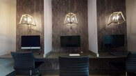 This thoughtfully designed single-floor amenity space combines contemporary style with practical functionality. The central lounge area serves as the heart of the space, featuring plush teal barrel chairs arranged around a round dark wood coffee table, set against a striking quilted teal accent wall adorned with a wall-mounted flat-screen TV and globe sconce lighting. A sputnik chandelier overhead adds a modern, artistic touch to the seating area.

Large floor-to-ceiling windows flood the space with natural light, offering views of the surrounding outdoor landscape. Warm linen drapes frame the windows, complementing the light ceramic tile flooring that runs throughout. A secondary seating nook near the entrance features a classic wingback chair, tufted ottomans, and a floor lamp, positioned beside tall windows for a cozy, well-lit reading corner.

A bold geometric patterned double door serves as a dramatic architectural focal point, while curated wall art adds visual character to the connecting hallway. The business center offers dedicated workstations with desktop computers, set beneath cage-style pendant lights against a richly textured dark stone-look wall, creating an atmosphere that is both productive and sophisticated. Clean corridors with dark-trimmed doors connect the various areas seamlessly.