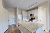 Located at 1111 Oakley Industrial Blvd in Fairburn, GA, this single-floor, one-bedroom, one-bathroom residence offers a thoughtfully designed layout that maximizes its compact footprint. The open-concept kitchen serves as the centerpiece of the main living area, showcasing a granite-topped island with a stainless steel sink, white shaker-style cabinetry, a tile backsplash, and a full suite of stainless steel appliances including a refrigerator, range, and microwave. Track lighting overhead adds a modern touch. Hardwood-style flooring flows through the kitchen and living area, while large windows and a glass door at the far end of the living space invite natural light and provide outdoor access. A ceiling fan enhances comfort in the main living area. The bedroom is carpeted for warmth and comfort, featuring two windows with views of the surrounding greenery and its own ceiling fan. The bathroom is finished with a granite vanity, white cabinetry, tile flooring, and a large mirror with vanity lighting. Directly adjacent to the bathroom is a dedicated walk-in closet with wire shelving for organized storage. A stacked in-unit washer and dryer is also conveniently included.
