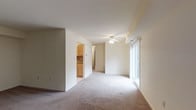 This single-floor space offers an open and airy layout with a clean, neutral aesthetic throughout. The main living area is finished with wall-to-wall carpet in a light beige tone, contributing to a warm and comfortable atmosphere. A ceiling fan is centrally mounted, providing both ventilation and subtle overhead lighting. Along one wall, a sliding glass door allows for an abundance of natural light to fill the room, brightening the open space and offering a connection to the outdoors.

The living area flows naturally toward an adjoining kitchen, visible through a defined opening in the wall. The kitchen features light wood cabinetry paired with tile flooring, creating a practical and inviting workspace that contrasts pleasantly with the carpeted living area. A tile backsplash adds a decorative touch to the kitchen's design.

The neutral white walls throughout create a versatile backdrop, allowing the space to feel open and adaptable. A hallway visible from the living area suggests additional rooms beyond, extending the functionality of the overall layout. The space maintains a straightforward, well-organized design that emphasizes comfort and practicality.