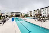 Located at 3616 N Rocky River Road in Monroe, North Carolina, this outdoor amenity space serves as a central gathering point for the surrounding residential community. The pool area is designed with a resort-style aesthetic, featuring a generously sized main swimming pool with clearly marked lap lanes for fitness use, alongside a freeform leisure pool that includes a distinctive circular wading island with Adirondack seating — ideal for relaxing in shallow water. The pool deck is finished in a light-toned, slip-resistant surface and is lined with rows of contemporary lounge chairs and side tables, providing ample space for sunbathing and outdoor socializing. A covered pavilion with a pitched roof anchors one end of the deck, offering shaded seating and a sheltered gathering area. The space is enclosed by a wrought iron perimeter fence, maintaining a sense of privacy and security. The multi-story residential buildings surrounding the courtyard feature a mix of white and brick exterior facades with balconies overlooking the pool area, creating an immersive community atmosphere. Lush greenery and landscaping accent the perimeter, softening the hardscape and adding visual appeal to this well-appointed outdoor recreational space.