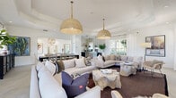 Located in Bradenton, Florida, this single-story space presents a cohesive, coastal-inspired design defined by crisp white walls, light-toned hardwood flooring, and an abundance of natural light streaming through large windows and glass-paned doors throughout.

The entry foyer makes an immediate impression with soaring ceilings, a striking geometric gold pendant light, and a tufted chaise lounge positioned beneath bold botanical artwork. Glass doors open directly to an outdoor pool area, establishing an effortless indoor-outdoor connection.

The expansive main gathering area features a tray ceiling with oversized woven dome pendant lights suspended above generously arranged seating. A central fireplace flanked by a wall-mounted television serves as the focal point, while sheer white drapery softens the surrounding windows and frames views of lush exterior greenery.

An open dining area adjoins the living space, furnished with a dark wood table, woven rattan chairs, and a decorative display cabinet. This zone transitions naturally into a compact, well-appointed kitchenette featuring dark navy cabinetry, white subway tile, a beverage refrigerator, and a dedicated coffee station.

A quieter sitting nook offers an intimate retreat with bright white walls, multi-pane windows, and curated artwork. Throughout, the overall ambiance is airy, refined, and relaxed.