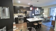 This contemporary single-floor space is designed with an open-concept layout that seamlessly connects the kitchen, dining, and living areas into one cohesive environment. The kitchen is a standout feature, boasting dark espresso cabinetry that extends to the ceiling, a crisp white countertop, and a full suite of stainless steel appliances including a gas range, built-in microwave, and a French door refrigerator with an ice and water dispenser. A central island with a white countertop provides additional prep space and casual seating with stylish industrial-style bar stools beneath. Pendant lighting hangs above the island, adding warmth and a modern aesthetic. The hardwood-style flooring runs throughout, lending continuity and a warm, grounded feel to the space. The kitchen transitions naturally into the living area, which is anchored by a neutral-toned sofa and ambient floor lamps, creating a relaxed and inviting atmosphere. An accent wall featuring bold, graphic wall art adds a touch of contemporary character. Recessed lighting throughout ensures the space is well-lit while maintaining a clean, uncluttered ceiling line. Overall, the space strikes a balance between modern design and functional living.