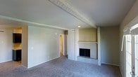 This single-floor space offers a welcoming and functional layout with thoughtfully chosen architectural details. The living area serves as the heart of the home, anchored by a classic brick fireplace framed by vertical shiplap-style wood paneling, creating a warm and timeless focal point. Exposed ceiling beams add a touch of rustic character to the otherwise clean, neutral aesthetic. The room is finished with plush wall-to-wall carpeting in a soft gray tone, contributing to a comfortable and quiet atmosphere. Recessed lighting complements the natural light filtering into the space, keeping the interior bright and inviting. The neutral gray and white color palette throughout the walls and trim provides a versatile backdrop suitable for a variety of interior styles. A hallway visible from the main living area leads to additional rooms, hinting at a practical and connected floor plan. The overall ambiance of the space is calm and understated, with its blend of traditional elements — such as the brick fireplace and wood beams — balanced by clean, modern finishes that give the home a timeless and move-in-ready appeal.