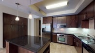 This thoughtfully designed single-floor residence offers a refined living experience through its consistent use of rich finishes and modern fixtures. The kitchen is a standout feature, showcasing dark espresso cabinetry, dark granite countertops, a subway tile backsplash, and a full suite of stainless steel appliances, including a French door refrigerator with water dispenser, built-in microwave, wall oven, and dishwasher. A central island with pendant lighting adds both workspace and visual interest, while recessed tray ceilings lend an architectural touch. The living areas are finished with warm hardwood flooring that flows throughout, complemented by large windows that invite generous natural light and views of the surrounding neighborhood. The bathroom features travertine-tiled walls surrounding a deep soaking tub, paired with dark wood cabinetry for storage. A dedicated laundry room provides full-size side-by-side washer and dryer units, dark cabinetry with a utility sink, and open shelving for added convenience. Spacious closets with double hanging rods and shelving offer ample storage. The consistent palette of dark wood tones, stone surfaces, and brushed stainless accents creates a cohesive, upscale ambiance throughout the space.