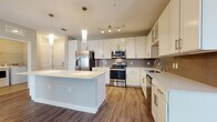 This single-story residence at 5671 Mauna Loa Blvd in Sarasota, Florida offers a thoughtfully designed open-concept layout with contemporary finishes throughout. The kitchen serves as the heart of the home, showcasing crisp white cabinetry with brushed hardware, a striking quartz island with pendant lighting overhead, a textured tile backsplash, and a full suite of stainless steel appliances including a range, microwave, refrigerator, and dishwasher. Wood-look flooring extends seamlessly from the kitchen into the open living and dining areas, which are brightened by natural light streaming through large windows and sliding glass doors. A ceiling fan and track lighting provide versatile illumination throughout the main living space. The bedrooms feature plush carpet, ceiling fans, and generous closet space, including a spacious walk-in closet with multi-level wire shelving. The bathrooms are finished with neutral tile flooring, white subway tile tub-shower surrounds, and vanities with quartz countertops. A dedicated laundry area houses a full-size GE washer and dryer. The overall ambiance is clean, bright, and move-in ready, reflecting a modern aesthetic well-suited to Florida living.