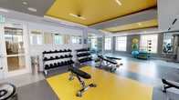 This contemporary fitness center is designed to support a full range of workout routines in a bright, energizing environment. The space is immediately defined by its striking architectural detail: bold yellow ceiling panels set against a gray and white palette, creating a vibrant, motivating atmosphere throughout. Linear LED lighting complements the natural light that streams in through a generous bank of windows, offering views of the surrounding greenery and keeping the space feeling open and airy.

The flooring features a dynamic combination of yellow and gray rubber tiles, delineating different workout zones while providing both durability and safety underfoot. The free weight area is well-stocked with a full rack of dumbbells and a set of kettlebells in varying weights, accompanied by adjustable benches for strength training exercises. Floor-to-ceiling mirrors along one wall visually expand the space and allow users to monitor their form during workouts.

The cardio section houses multiple treadmills, an elliptical trainer, a stationary upright bike, and dedicated spin bikes, all positioned to take advantage of the outdoor views. A functional training zone features stability and medicine balls for dynamic exercises. The space is rounded out by a cable machine and additional strength equipment, making this fitness center a comprehensive facility suited for users of all fitness levels.