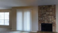 This single-floor residence offers a welcoming and practical layout designed for comfortable everyday living. The living area serves as the heart of the home, immediately drawing attention with its striking floor-to-ceiling stone fireplace — a standout architectural feature that adds rustic character and warmth to the space. The natural stonework is rich in texture and color, creating a focal point that anchors the room with timeless charm.

A large sliding glass door dressed with vertical blinds allows for adjustable natural light and provides a seamless connection to the outdoors. When the blinds are drawn open, the space is flooded with daylight, enhancing the open and airy feel of the interior. The neutral-toned walls and carpeted flooring contribute to a soft, understated palette that complements the earthy tones of the stonework.

A textured popcorn ceiling adds a classic finish overhead, consistent with the home's traditional character. The overall ambiance of the space is warm and inviting, with a balance of functional simplicity and cozy architectural detail. This single-level layout offers ease of movement throughout, making it a practical and comfortable place to call home.