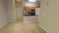 This single-floor residence offers an open-concept design that connects the kitchen and living areas into one cohesive, functional space. The kitchen is well-appointed with warm honey-toned wood cabinetry, dark granite countertops, and a full suite of stainless steel appliances including a refrigerator, microwave, and gas range. A peninsula counter extends into the living space, providing additional workspace and a natural division between the kitchen and the main living area without interrupting the open feel. The flooring transitions from easy-care surfaces near the entry and kitchen to soft, neutral-toned wall-to-wall carpeting throughout the living area, adding warmth underfoot. The entry foyer is defined by a solid front door with a security deadbolt, offering a welcoming arrival point. Neutral beige walls throughout create a clean, versatile backdrop that suits a variety of furnishing styles. Overhead lighting keeps the space well-illuminated, and a window in the kitchen area allows for natural light. The overall layout is practical and efficiently designed, making thoughtful use of its footprint to create a comfortable and inviting living environment.