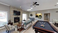 This single-floor community clubhouse offers a thoughtfully designed space for socializing, recreation, and relaxation. The heart of the space is a generously sized open lounge area finished with light wood-look flooring, crown molding, and a textured ceiling. A striking stacked-stone accent wall serves as the focal point, housing a fireplace beneath a wall-mounted flat-screen television, creating a warm and inviting atmosphere. Comfortable seating arrangements, including a plush sofa draped with a cozy throw, accent chairs, and area rugs, complete the living area. A full-size pool table anchors the recreational side of the lounge, adding an engaging entertainment element.

An open pass-through connects the lounge to a kitchenette area, providing convenient access for gatherings. Adjacent to the main lounge is a casual dining and activity nook featuring a round high-top table with stools, board games, a water station, and a computer workstation — ideal for both leisure and productivity. Large glass French doors lead from a secondary seating area into a dedicated fitness center, which is equipped with cardio machines and a multi-use cable gym system. Natural light flows throughout via multiple windows, enhancing the open, welcoming ambiance of the entire space.