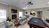 This single-floor community clubhouse offers a thoughtfully designed space for socializing, recreation, and relaxation. The heart of the space is a generously sized open lounge area finished with light wood-look flooring, crown molding, and a textured ceiling. A striking stacked-stone accent wall serves as the focal point, housing a fireplace beneath a wall-mounted flat-screen television, creating a warm and inviting atmosphere. Comfortable seating arrangements, including a plush sofa draped with a cozy throw, accent chairs, and area rugs, complete the living area. A full-size pool table anchors the recreational side of the lounge, adding an engaging entertainment element.

An open pass-through connects the lounge to a kitchenette area, providing convenient access for gatherings. Adjacent to the main lounge is a casual dining and activity nook featuring a round high-top table with stools, board games, a water station, and a computer workstation — ideal for both leisure and productivity. Large glass French doors lead from a secondary seating area into a dedicated fitness center, which is equipped with cardio machines and a multi-use cable gym system. Natural light flows throughout via multiple windows, enhancing the open, welcoming ambiance of the entire space.