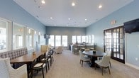 This single-floor communal space is designed with comfort and functionality in mind, offering a versatile gathering area that balances open-plan living with defined seating zones. The room is finished in soft, muted blue tones that lend a calming and sophisticated ambiance throughout.

The space features a variety of seating arrangements to accommodate different group sizes and occasions. A circular dining table with upholstered chairs anchors one side of the room, while a long wooden communal table with cross-back chairs lines the adjacent wall. A cozy lounge nook furnished with plush cream armchairs provides a more relaxed setting for informal conversations.

At the far end of the room, a kitchenette-style bar counter with seating offers a convenient social hub, complemented by pendant lighting overhead. Recessed ceiling lighting is distributed evenly throughout, while large windows along the back wall invite generous amounts of natural light into the space, keeping the interior bright and airy.

The neutral carpeted flooring ties the room together seamlessly, and a wall-mounted flat-screen television adds a practical media element. Dark-framed glass panel doors provide an elegant transition point, enhancing the overall refined aesthetic of this well-appointed communal lounge.