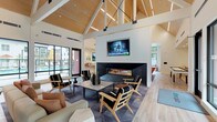 This single-story communal space showcases a striking architectural design anchored by soaring vaulted ceilings clad in warm natural wood paneling, supported by exposed white structural beams that lend the interior a contemporary barn-inspired aesthetic. Integrated linear lighting runs along the ceiling, complementing abundant natural light that pours in through large black-framed windows and glass doors, offering views of the surrounding outdoor areas.

The main lounge is thoughtfully divided into distinct seating zones. One area centers on a sleek, floor-to-ceiling linear gas fireplace with a dark tile surround, mounted flat-screen television, and an arrangement of black-and-white accent chairs paired with a white coffee table. An adjacent zone features bold orange upholstered chairs grouped around round metal side tables atop a geometric-patterned area rug, creating a vibrant social space. A wall-mounted television and bar-height counter with seating further enhance this area.

Toward the rear, a billiards table sits beneath expansive windows that frame outdoor greenery. The kitchenette is outfitted with wood-grain cabinetry, light quartz countertops, stainless steel appliances including a glass-door refrigerator and dishwasher. Light wood-look flooring flows throughout, unifying the space with a clean, modern finish.