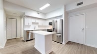 This single-floor unit presents a clean, contemporary design characterized by a neutral palette of whites and soft grays accented by light wood-look flooring that flows consistently throughout the space. The open-concept kitchen serves as a central highlight, outfitted with gray shaker-style cabinetry, white quartz countertops, a white subway tile backsplash, and a full suite of stainless steel appliances including a French door refrigerator, range, microwave, and dishwasher. A freestanding center island with a white quartz surface provides additional prep space and a natural visual anchor between the kitchen and the generously sized living area. Recessed and pendant lighting enhance the bright, well-lit atmosphere throughout.

The bedrooms are finished in the same light wood-look flooring and feature neutral walls, ample natural light from windows with blinds, and access to closet storage. Two bathrooms serve the unit; one features gray vanity cabinetry, a backlit mirror, white quartz countertop, and a tub-shower combination with white tile surround, while the second offers a similarly clean and functional design. Overall, the space conveys a modern, move-in-ready aesthetic with consistent finishes and a cohesive, understated style.