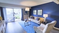 This single-floor space centers around a thoughtfully designed living room that makes a strong visual statement through its use of color and contemporary furnishings. The most defining feature is a deep navy blue accent wall, which serves as a dramatic backdrop for two framed art pieces and anchors the room's bold aesthetic. The remaining walls are finished in a soft neutral tone, creating a balanced contrast that prevents the space from feeling overwhelming.

Hardwood-style flooring runs throughout, lending warmth and cohesion to the open layout. A chevron-patterned blue area rug defines the seating area and reinforces the room's color palette. The living space is furnished with a neutral sofa accented by blue and yellow throw pillows, alongside a textured accent chair and sleek nesting coffee tables.

Natural light is a notable feature, streaming in through large sliding glass doors fitted with patterned drapes. These doors open directly to an outdoor patio area, where lush greenery is visible, offering a pleasant indoor-outdoor connection. Overhead, a contemporary drum pendant light provides additional illumination, while a textured ceiling adds subtle architectural character to the room.