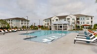 Located at 7301 Topwater Dr in Wilmington, North Carolina, this outdoor amenity space offers a resort-inspired pool experience within a well-appointed residential community. The centerpiece is a generously sized swimming pool with crystal-clear blue water, featuring gradual entry points and in-water lounge ledges that invite relaxation. Surrounding the pool, a spacious concrete sun deck is lined with cushioned chaise lounges accented with vibrant orange pillows, providing ample space for sunbathing. Two covered pavilion structures anchor opposite ends of the pool area. One serves as an open-air cabana lounge, furnished with comfortable seating, a wall-mounted outdoor television, and a hanging chair, making it ideal for social gatherings. The other features a metal roof and serves as an additional shaded retreat. Landscaping throughout the space includes neatly manicured ornamental grasses, mulched garden beds, and tall palm trees that reinforce the coastal Carolina atmosphere. An adjacent outdoor courtyard offers shaded dining tables beneath large rust-colored umbrellas, surrounded by lush greenery. Black wrought-iron fencing encloses the entire area, adding security while maintaining an open, airy feel. The multi-story residential buildings visible in the background feature white and gray coastal architectural details with metal roofing.
