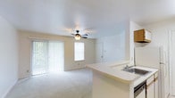 This single-floor residence offers a functional and open layout designed to maximize livability. The main living area is carpeted throughout and centers around a ceiling fan with integrated lighting, providing both comfort and illumination. A large sliding glass door with vertical blinds lines one wall, drawing in abundant natural light and offering a view of the outdoors, while an additional window further brightens the space.

The kitchen connects seamlessly to the living area via a built-in peninsula counter, which includes an integrated sink and extends the workspace while maintaining an open, connected feel. White cabinetry and light-toned countertops keep the kitchen area clean and bright. Ventilation registers are visible at floor level, indicating a built-in climate control system throughout. Neutral white walls and light-colored carpet unify the space, creating a clean and airy atmosphere throughout. A hallway leads to additional rooms, including what appears to be a closet or utility space, suggesting practical storage solutions. Overall, the layout is straightforward and well-organized, with a consistent, understated aesthetic that offers a versatile canvas for personalization.