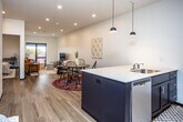 This well-designed single-floor unit offers a clean, modern aesthetic anchored by an open-concept layout that seamlessly connects the kitchen, dining, and living areas. The kitchen is a standout feature, equipped with white shaker cabinetry, stainless steel appliances including a refrigerator, range, and microwave, and a striking dark-finished island with a white countertop, an integrated sink, and bar seating. A deep blue tile backsplash adds a bold accent against the bright cabinetry, while stylish pendant lights hang overhead. Recessed lighting throughout the main living area ensures a bright, inviting atmosphere. The open living and dining space is generously proportioned, with warm wood-look flooring and large windows that draw in natural light and provide outdoor views. Two bedrooms offer comfortable accommodations; one features a wall-mounted mini-split HVAC unit and a sleek barn-style sliding door, while the other includes a window with a pleasant exterior view and space for a work desk. The bathroom is finished with light tile flooring, a dark vanity with a white countertop, and clean white walls. Throughout the unit, a cohesive palette of white walls, dark accents, and natural wood tones creates a contemporary and functional living environment.