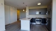 This single-floor space showcases a contemporary open-concept layout anchored by a well-appointed kitchen. The kitchen features two-tone cabinetry, combining dark lower cabinets with grey upper cabinets that extend toward the ceiling, maximizing storage capacity. A striking white waterfall island serves as the centerpiece, equipped with an undermount sink and a modern gooseneck faucet. Two pendant lights hang above the island, providing focused task lighting that complements the flush ceiling fixture overhead.

Stainless steel appliances, including a built-in microwave, electric range, and dishwasher, lend a polished, professional quality to the kitchen. The light-colored countertops contrast cleanly against the dark cabinetry, reinforcing the modern aesthetic throughout.

Medium-toned hardwood-style flooring runs continuously through the kitchen and into the adjacent living area, creating a cohesive visual flow. A track lighting system along the ceiling of the adjoining hallway ensures the space remains well-lit. A neutral color palette of whites and greys throughout the space contributes to a clean, minimalist ambiance. A hallway visible in the background leads to additional areas of the unit.