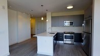 This single-floor space showcases a contemporary open-concept layout anchored by a well-appointed kitchen. The kitchen features two-tone cabinetry, combining dark lower cabinets with grey upper cabinets that extend toward the ceiling, maximizing storage capacity. A striking white waterfall island serves as the centerpiece, equipped with an undermount sink and a modern gooseneck faucet. Two pendant lights hang above the island, providing focused task lighting that complements the flush ceiling fixture overhead.

Stainless steel appliances, including a built-in microwave, electric range, and dishwasher, lend a polished, professional quality to the kitchen. The light-colored countertops contrast cleanly against the dark cabinetry, reinforcing the modern aesthetic throughout.

Medium-toned hardwood-style flooring runs continuously through the kitchen and into the adjacent living area, creating a cohesive visual flow. A track lighting system along the ceiling of the adjoining hallway ensures the space remains well-lit. A neutral color palette of whites and greys throughout the space contributes to a clean, minimalist ambiance. A hallway visible in the background leads to additional areas of the unit.