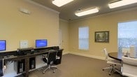 This single-floor space is designed with functionality and practicality in mind, offering a well-appointed work environment suited for focused productivity. The room features warm, neutral-toned walls complemented by crisp white crown molding, lending a polished and professional aesthetic throughout. Carpeted flooring adds a layer of comfort underfoot, while bright flush-mount ceiling fixtures provide ample, even illumination across the entire space. A dedicated workstation area is set up with a dark-surfaced desk, a desktop computer, and an ergonomic office chair, creating an efficient setup for computer-based tasks. An additional chair is also present, offering flexible seating for collaboration or guests. A window fitted with horizontal blinds allows for natural light control, keeping the ambiance comfortable throughout the day. A framed landscape painting adorns one wall, adding a subtle decorative touch that softens the utilitarian nature of the room. The layout is open and uncluttered, maximizing usability and ease of movement. Overall, this space delivers a clean, organized, and inviting environment well-suited for office work or business use.