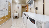 Located at 145 Jameson Ave in Toronto's Parkdale neighbourhood, this shared laundry facility is a compact, well-organized space designed for practical, everyday use. The room is finished with cream-toned ceramic wall tiles from floor to ceiling, lending a clean and hygienic aesthetic throughout. Durable terrazzo flooring provides a hard-wearing surface suited to high-traffic use.

The facility is equipped with a row of commercial-grade Speed Queen washers and dryers, offering residents reliable and efficient laundry capabilities. Flexible ventilation ductwork runs overhead from the dryer units, ensuring proper airflow. A standalone utility sink with exposed plumbing is also present, adding functional versatility to the space.

Fluorescent overhead lighting illuminates the room evenly, while posted instructional signage and usage guidelines are clearly displayed on the walls for resident reference. An adjacent corridor connects to a service room, providing additional utility access. The space also includes waste bins and basic cleaning equipment, supporting general upkeep of the facility.

Overall, this laundry room is a straightforward, efficiently arranged shared amenity that prioritizes function and cleanliness, serving as a practical resource for residents of the building.