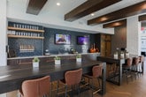 Located on Bell Road in Johns Creek, Georgia, this single-floor amenity space combines contemporary design with a warm, social atmosphere. The layout is open and thoughtfully organized into distinct functional zones, all unified by striking herringbone-patterned hardwood flooring and exposed dark wood beam accents overhead.

The bar and lounge area serves as a central focal point, featuring rich walnut-toned cabinetry with brushed gold hardware, a light quartz countertop, a stainless undermount sink, and a dramatic blue-gray glazed tile backsplash. Floating oak shelves display glassware, and mounted flat-screen televisions make this area well-suited for social gatherings. A draft beer station adds a hospitality-forward touch.

Adjacent seating areas include both high-top communal tables with leather counter chairs and a more intimate round dining arrangement beneath a pendant light, surrounded by velvet upholstered chairs. Curated abstract artwork adorns the walls alongside white-painted brick accents that lend an industrial warmth to the space.

Floor-to-ceiling black-framed windows and glass walls flood the interior with natural light and frame views of the surrounding wooded landscape, creating an inviting, light-filled environment throughout.