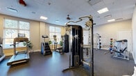 This fitness center is organized across a single floor and divided into two distinct exercise rooms, offering a comprehensive range of workout options. The main exercise room is spacious and thoughtfully outfitted, featuring rubber flooring throughout for safety and durability. It houses multiple treadmills, elliptical machines, a stair climber, stationary bikes, and a full rack of dumbbells positioned in front of a large wall mirror that enhances the sense of space and allows for form-checking. A multi-functional cable machine and a heavy punching bag anchor the strength training area, accompanied by a squat rack, weight bench, barbells, and a medicine ball rack. Natural light fills the room through multiple windows fitted with blinds, and ceiling fans contribute to airflow and comfort. Recessed panel lighting in the drop ceiling maintains a bright, clean atmosphere. The second exercise room offers a more open, flexible environment with a cushioned cork-style floor, two wall-mounted flat-screen televisions, stability balls, and a plyometric box — ideal for group classes, stretching, or functional training. This room also benefits from natural light via a tall window with outdoor views. Together, the two rooms provide a well-rounded fitness facility suitable for a wide variety of training styles.