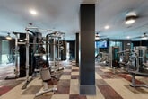 This single-level fitness center is designed with a clear emphasis on both functionality and contemporary style. The space is organized into distinct zones, each accessible via a sleek corridor lined with white walls, gold-accented wall sconces, and large-format tile flooring. Signage identifies dedicated "Strength" and "Tone" areas, framed by floor-to-ceiling glass partitions with matte black steel frames that lend the facility a modern, upscale aesthetic.

The strength training area features a checkerboard-patterned tile floor in earthy tones and is outfitted with cable machines, weight benches, a Smith machine, a punching bag, and free weights including a full rack of colorful kettlebells. Dark accent walls and ceiling fans with integrated lighting contribute to an energetic yet polished atmosphere. Mounted flat-screen televisions are positioned throughout for entertainment during workouts.

The cardio zone offers treadmills, ellipticals, and stair climbers positioned near large windows that frame outdoor views, filling the space with natural light. Full-length mirrors amplify the sense of openness throughout.

A separate studio area provides barre rails, wall-to-wall mirrors, branded yoga mats, and patterned carpet flooring, making it well-suited for group fitness and mind-body training sessions.