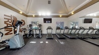 Located at 70 Mowry Ave in Fremont, California, this fitness center offers a comprehensive workout environment within a single-story space designed with both functionality and aesthetic appeal in mind. The room features a striking tray ceiling with crown molding and a warm gold accent band, lending an elevated, polished character uncommon in typical gym settings. Recessed and wall-mounted lighting complements the natural light that filters in through multiple windows fitted with blinds, creating a bright and energizing atmosphere throughout the day.

The cardio zone is well-appointed with multiple treadmills and elliptical trainers arranged along one wall, accompanied by a wall-mounted flat-screen television for entertainment during workouts. A full-length mirror along one wall visually expands the space and aids in form monitoring. The strength training area features a multi-station cable machine, an adjustable bench, and a rack of dumbbells, providing a range of resistance training options. Additional equipment includes upright and recumbent bikes, a stair climber, and yoga accessories stored along the walls. A built-in cubby storage unit keeps the space organized. Bold "Train Hard" wall graphics add a motivational touch. Double French doors open directly to an outdoor pool area, enhancing ventilation and outdoor connectivity.