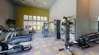 Located at 722 Roper Pond Cir in Columbia, SC, this fitness center is a well-equipped, single-floor facility designed to support a comprehensive workout experience. The space features soaring ceilings that create an open, airy atmosphere, complemented by an abundance of natural light streaming through large multi-pane windows that offer views of the surrounding greenery and outdoor pool area.

The flooring throughout consists of a distinctive grey and yellow patterned carpet tile, providing both durability and visual character. Floor-to-ceiling mirrors line one wall, enhancing the sense of space and allowing members to monitor their form during exercise.

The cardio zone is outfitted with treadmills, elliptical trainers, and stationary spin bikes, all arranged to take advantage of the natural light and outdoor views. A dedicated strength training area features a multi-station cable machine suitable for a wide range of resistance exercises. Free weights are neatly organized on a tiered dumbbell rack along the mirrored wall, offering a full range of weight options.

Additional accessories including balance equipment, exercise mats, and weight benches are available throughout the space. Wall-mounted televisions provide entertainment during workouts. Recessed lighting and a clean, neutral color palette with accent walls in warm olive green round out the inviting, functional environment.