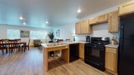 This single-floor space features an open-concept layout that seamlessly connects the kitchen, dining area, and living room, creating a cohesive and functional environment. Light oak hardwood-style flooring runs throughout, complemented by white walls and recessed lighting that provide a clean, bright atmosphere.

The kitchen is well-appointed with light wood cabinetry offering generous storage, black appliances including an electric range, refrigerator, and dishwasher, and a peninsula island featuring a double sink and bar seating. The light-toned countertops contrast neatly with the dark appliances, giving the kitchen a practical yet polished character.

Adjacent to the kitchen, the dining area accommodates a large dark wood table with seating for up to eight, positioned near windows that allow natural light to fill the space. A wall-mounted flat-screen television is also present in this area.

The living room is furnished with a leather loveseat, upholstered armchair, and a glass-top coffee table. Large windows with horizontal blinds allow ample natural light while providing privacy. Abstract wall art and decorative plants add warmth and visual interest to the overall space.