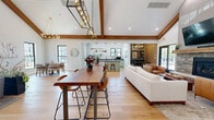 Located at 103 Laurel Hill Rd in Brookfield, Connecticut, this thoughtfully designed single-floor space combines modern farmhouse aesthetics with warm, inviting details throughout. Soaring vaulted ceilings with rich, exposed wood beams define the architectural character of the space, creating an airy, open atmosphere enhanced by wide-plank hardwood flooring that flows seamlessly from room to room.

The living room serves as a central gathering point, anchored by a striking stone fireplace with a rustic wood mantel and a wall-mounted television above. Comfortable seating, including plush sofas and leather accent chairs, is arranged around a woven area rug, while glass doors lead directly to an outdoor patio, inviting natural light and views of the surrounding greenery.

The adjacent dining area features a dramatic circular chandelier suspended from the vaulted ceiling, illuminating a round dining table surrounded by modern upholstered chairs. Large black-framed windows frame views of the exterior, flooding the area with natural light.

The kitchen is a standout with sage green cabinetry, dark countertops, a subway tile backsplash, and a central island with bar seating. Statement pendant lights and nautical-inspired décor add personality and charm.

A cozy sitting nook with artisan wood shelving, framed artwork, and accent chairs rounds out this well-appointed space, offering a quiet retreat within the larger layout.
