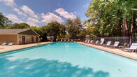 Located at 1331 Wicklow Dr in Cary, NC, this outdoor amenity space centers around a generously sized swimming pool with clear, vibrant turquoise water. The pool is rectangular in shape and accommodates both leisurely swimming and relaxation, with entry points accessible from a broad surrounding concrete sun deck. The deck is well-furnished with rows of lounge chairs positioned along the poolside, offering ample space for sunbathing and unwinding. Several umbrella-shaded seating areas provide relief from direct sun, featuring blue sling-style lounge chairs and side tables ideal for outdoor lounging.

The entire pool area is enclosed by a black wrought iron fence, providing both security and an open, airy feel that maintains visibility to the surrounding landscape. Beyond the fence, mature deciduous and evergreen trees create a dense, natural backdrop that lends the space a private, serene atmosphere. Mulched landscaping beds with low shrubs line the perimeter, contributing to the well-maintained, park-like setting.

A small accessory structure — likely a utility or storage building — is visible at the edge of the property. The open sky above, combined with the towering tree canopy, gives the space a refreshing outdoor ambiance well-suited for warm-weather enjoyment.