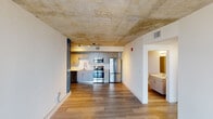 Located on the penthouse level at 35 SW 6th Ave in Miami's vibrant Brickell area, unit PH1 is a single-floor residence that blends industrial design elements with contemporary finishes. The unit is accessed via a dedicated penthouse elevator lobby, accented by a bold red feature wall and large-format tile flooring, opening onto a wide, wood-floored corridor with a distinctive exposed raw concrete ceiling that carries throughout the interior.

The open-concept living area is defined by its striking exposed concrete ceiling and light-toned wood plank flooring. A deep navy accent wall adds a dramatic design element, while expansive floor-to-ceiling windows fill the space with natural light and frame sweeping panoramic views of the Miami skyline and surrounding cityscape.

The kitchen is well-appointed with sleek gray textured cabinetry extending to the ceiling, quartz countertops, a stainless steel single-basin sink with an industrial-style faucet, and a full suite of stainless steel appliances including a French door refrigerator, electric range with oven, and built-in microwave. Track lighting on the exposed concrete ceiling provides focused task illumination.

A bedroom features the same raw concrete ceiling and includes a wide double-panel closet with louvered doors, offering ample storage. A bathroom with vanity cabinetry is conveniently accessible from the main living area. Throughout the unit, clean white walls contrast with the raw structural ceiling to create a modern industrial aesthetic.
