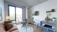 This single-floor space showcases a clean, modern design with an open-concept layout that seamlessly connects the living and dining areas. The most striking feature is the expansive floor-to-ceiling window along one wall, which floods the interior with abundant natural light and offers elevated views of a leafy, residential neighborhood below.

The living area is furnished with a warm-toned sofa and an accent chair, centered around a round glass coffee table atop a circular area rug, creating an inviting and functional seating arrangement. A wall-mounted flat-screen television is positioned across from the seating area, complemented by a compact desk and metal chair beneath it, providing a dedicated workspace.

Adjacent to the living area, a high-top dining table with matching stools offers casual dining. The light hardwood flooring runs throughout, enhancing the sense of openness and warmth. White walls and a neutral ceiling contribute to the airy, minimalist aesthetic. A vertical HVAC unit is integrated into the wall, maintaining functionality without disrupting the overall clean design. The space strikes a balance between contemporary style and everyday practicality.