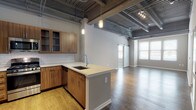 This single-floor loft-style space showcases a compelling blend of industrial and contemporary design elements. The most striking architectural feature is the dramatically high ceiling, left intentionally exposed to reveal corrugated metal decking, structural beams, ductwork, and piping — all finished in dark tones that reinforce the industrial aesthetic throughout.

The open-concept kitchen is a central highlight, featuring warm wood-grain cabinetry with sleek hardware, white quartz countertops, and a classic white subway tile backsplash. A peninsula with an undermount stainless steel sink and pull-down faucet extends into the living area, creating a natural gathering point. A full suite of stainless steel appliances — including a gas range, built-in microwave, dishwasher, and refrigerator — completes the well-equipped kitchen.

The expansive living area flows seamlessly from the kitchen, with rich hardwood flooring underfoot and a white sliding barn door that adds a distinctive architectural touch. Multiple windows allow generous natural light to fill the space. A bedroom features plush carpet and its own window, while the bathroom offers a wood-grain vanity, white quartz countertop, and a combination tub and shower. Pendant lighting throughout adds warmth to the otherwise industrial palette.
