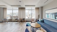 Located at 1100 2nd Place SE in Washington DC, this thoughtfully designed hospitality suite delivers a contemporary aesthetic with a bold, colorful palette throughout. The main lounge area serves as the heart of the space, anchored by a deep blue sofa adorned with teal, yellow, and multicolored accent pillows, paired with a round wood-top coffee table and a vibrant geometric area rug. Curated wall art and recessed lighting add to the polished atmosphere, while large floor-to-ceiling windows flood the room with natural light and offer expansive views of the Washington DC skyline and surrounding cityscape.

A dedicated dining and lounge nook with round bistro-style tables and contemporary seating provides a relaxed setting for casual gatherings. The bar area is a standout feature, showcasing a striking teal subway-tile backsplash, pendant lighting, a long white countertop with built-in power access, teal barstools, and integrated appliances including beverage refrigerators and mounted screens. A formal conference room rounds out the suite, featuring a substantial walnut dining table, upholstered seating, an elegant layered chandelier, and dramatic floor-to-ceiling windows that frame sweeping views of the city below. The overall ambiance is modern, energetic, and well-appointed for both professional and social occasions.