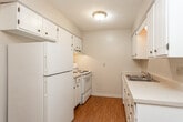 This single-story unit offers a straightforward and functional layout designed for everyday living. The kitchen is equipped with white cabinetry, a single-basin sink with light countertops, a white refrigerator, and an electric range. Wood-look flooring runs throughout the kitchen area, lending a warm contrast to the white finishes. An overhead ceiling fan with pendant lighting serves the adjoining dining or eating area, which flows naturally from the kitchen space.

The living area is carpeted in a neutral beige tone and features a front entry door, providing a welcoming transition into the home. Natural light enters through windows fitted with blinds. The space connects visibly to the kitchen, creating an open and cohesive common area.

The bedrooms are finished with the same neutral carpet and painted in light, neutral tones. Each room features a window that allows in natural light and offers exterior views, contributing to an airy atmosphere. Textured ceilings are present throughout the carpeted rooms.

The bathroom includes a full tub-shower combination with large-format beige tile lining the surround, accented by a decorative mosaic border in black and metallic tones. Chrome fixtures and a white toilet complete the clean, modern-neutral aesthetic. Overall, the unit maintains a consistent, understated palette throughout.