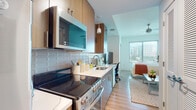 This contemporary studio presents a thoughtfully designed open-concept layout that seamlessly integrates living, sleeping, and kitchen functions within a single, well-organized space. The kitchen is outfitted with stainless steel appliances, including an electric range, over-the-range microwave, and refrigerator. Warm wood-grain cabinetry pairs with white quartz countertops and a subway tile backsplash, creating a clean, modern aesthetic. A pendant light fixture above adds a refined touch.

The living and sleeping area benefits from large floor-to-ceiling windows that invite substantial natural light and offer open urban views of the surrounding cityscape. A ceiling fan with a modern profile services the space for added comfort. Hardwood-style flooring runs throughout, adding warmth and visual continuity from the kitchen through the main living area.

A hallway connects the main space to the full bathroom, which features a wood-grain vanity with a white countertop, an integrated sink, a large wall mirror, overhead cabinetry for storage, and a shower with a curtain enclosure. The overall design employs a neutral palette of whites and natural wood tones, creating a bright, cohesive, and functional living environment well-suited to modern urban living.