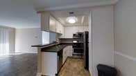 This single-floor space offers a thoughtfully designed layout with a focus on modern, functional living. The kitchen stands as a highlight, featuring crisp white shaker-style cabinetry that extends to the ceiling, accented by dark countertops that provide a striking contrast. A peninsula with an undermount sink and a built-in dishwasher offers additional workspace and the potential for casual dining or entertaining. The kitchen is equipped with a full suite of stainless steel appliances, including a gas range with double oven, an over-the-range microwave, and a black French door refrigerator, making it both practical and stylish. A tray ceiling with a flush-mount light fixture adds architectural interest and a sense of depth overhead. Warm wood-look flooring runs throughout, lending continuity and a welcoming ambiance to the space. Crown molding along the upper trim adds a refined finishing touch. The kitchen opens to an adjacent living area, allowing for a natural flow between spaces and enhancing the overall sense of openness. Natural light filters in from the neighboring room, contributing to a bright and inviting atmosphere throughout this well-crafted single-level home.