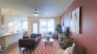 This single-story space showcases a thoughtfully designed living area that balances comfort and style. The living room is anchored by a striking deep-red accent wall, which adds warmth and visual depth to the space, complementing the neutral cream and beige tones found throughout. A bold, coral-patterned area rug grounds the seating arrangement and introduces an energetic design element to the room.

The space is furnished with a dark leather sofa and coordinating upholstered seating, accented with teal throw pillows that provide refreshing pops of color. A glass-top coffee table sits at the center, enhancing the open, airy feel of the room. A wall-mounted flat-screen television is positioned on a rustic media console along one wall, while large-scale wall art adds a decorative focal point nearby.

Natural light flows generously into the room through multiple windows dressed with white horizontal blinds and sheer curtains, creating a bright and inviting atmosphere. A ceiling fan with integrated lighting ensures year-round comfort. Lush indoor plants are placed throughout, adding organic texture and life to the interior. A connected kitchen area is partially visible, suggesting an open-concept floor plan.