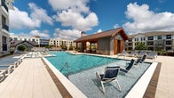 Located at 525 Sawnee Village Boulevard in Cumming, Georgia, this expansive outdoor amenity area is designed to deliver a resort-style experience. The centerpiece of the space is a generously sized main swimming pool with crystal-clear blue water, flanked by wide, light-toned tile pool decks lined with rows of cushioned lounge chairs. A second, shallower wading pool sits adjacent, providing a more relaxed water feature complete with in-pool seating. A standout architectural element is the rustic poolhouse pavilion, constructed with warm wood timber framing, a metal roof, and a striking stone fireplace chimney. This structure serves as a covered gathering and seating area, adding both function and visual character to the space. Elevated sun deck platforms with multi-tiered composite decking offer additional lounging areas with a distinct vantage point over the pools. The surrounding hardscape features patterned brick and stone pavers, while manicured landscaping with shrubs and trees provides natural accents throughout. The multi-story residential buildings that border the pool area feature stone and board-and-batten exterior details, large windows, and private balconies that overlook the amenity space. The entire area is enclosed by wrought iron fencing, ensuring a defined, secure environment open to the sky above.