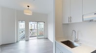 This contemporary single-floor space showcases a clean, minimalist aesthetic defined by an open-concept design that seamlessly connects the kitchen and living areas. The interior is bathed in natural light, enhanced by a crisp all-white color palette and light-toned hardwood-style flooring that runs throughout the space.

The kitchen is thoughtfully appointed with white shaker-style cabinetry featuring brushed nickel hardware, a stone-speckled composite countertop, and a deep stainless steel sink with a modern arc-neck faucet. Ample upper and lower cabinet storage ensures functional use of the space.

The living area is open and airy, anchored by a stylish drum pendant light fixture that adds a warm, inviting focal point. A full-width sliding glass door opens to a private balcony, offering views of the surrounding urban cityscape and filling the interior with abundant natural light throughout the day.

The balcony, enclosed by a sleek metal railing, provides an ideal outdoor retreat with a dynamic city backdrop. The overall ambiance of the space is modern, bright, and effortlessly functional, making it well-suited for those who appreciate contemporary urban living with a clean, uncluttered design sensibility.