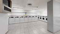 Located at 765 Steeles Ave W in Toronto, this shared laundry facility offers a clean, well-organized space designed for practical, everyday use. The room is finished with large-format white ceramic floor tiles that lend a bright, sanitary feel throughout. Overhead fluorescent lighting ensures the entire space is evenly and thoroughly illuminated.

The main laundry area houses a generous row of front-loading washing machines and dryers, all finished in white and lined along two walls, offering ample capacity for multiple users. The machines are clearly labeled and accompanied by posted instructional signage to assist users. A utility sink and a countertop surface provide additional functionality for laundry preparation.

A structural tiled column divides portions of the room, adding architectural definition while maintaining an open and navigable layout. An adjacent anteroom features a wall-mounted display screen, a waste receptacle, a notice board, and wall-mounted utility panels, providing an organized space for information and building services. A grey steel door connects this area to the rest of the building, including elevator access.

The facility's white walls, clean sightlines, and practical layout make it a highly functional communal amenity. The overall ambiance is orderly and efficient, reflecting a well-maintained shared-use environment suited to a residential building.