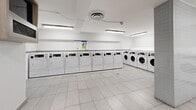 Located at 765 Steeles Ave W in Toronto, this shared laundry facility offers a clean, well-organized space designed for practical, everyday use. The room is finished with large-format white ceramic floor tiles that lend a bright, sanitary feel throughout. Overhead fluorescent lighting ensures the entire space is evenly and thoroughly illuminated.

The main laundry area houses a generous row of front-loading washing machines and dryers, all finished in white and lined along two walls, offering ample capacity for multiple users. The machines are clearly labeled and accompanied by posted instructional signage to assist users. A utility sink and a countertop surface provide additional functionality for laundry preparation.

A structural tiled column divides portions of the room, adding architectural definition while maintaining an open and navigable layout. An adjacent anteroom features a wall-mounted display screen, a waste receptacle, a notice board, and wall-mounted utility panels, providing an organized space for information and building services. A grey steel door connects this area to the rest of the building, including elevator access.

The facility's white walls, clean sightlines, and practical layout make it a highly functional communal amenity. The overall ambiance is orderly and efficient, reflecting a well-maintained shared-use environment suited to a residential building.